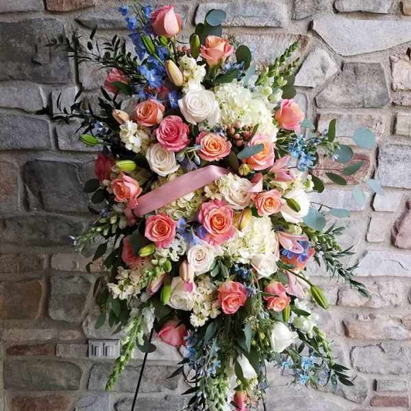 Tall standing spray of peach and white roses with blue accents on a metal easel against a stone wall.