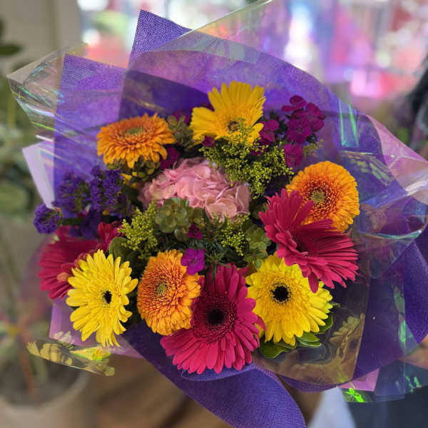 Bright hand-tied bouquet of pink, yellow, and orange gerbera daisies with mixed blooms in purple wrap