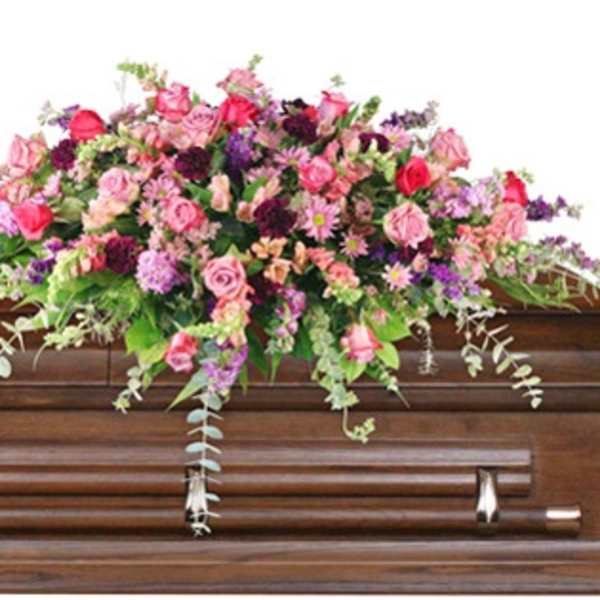 Pink and purple mixed flower casket spray in a lush, cascading style on a wooden casket