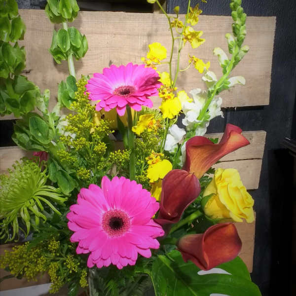 Mixed bouquet with pink gerbera daisies, yellow blooms, and burgundy calla lilies in a glass vase.