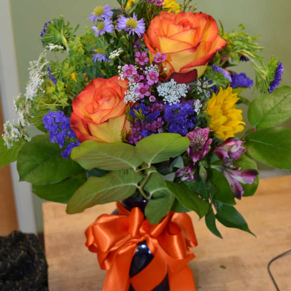 Bouquet of orange roses, purple flowers, and yellow blooms in a dark vase with an orange ribbon