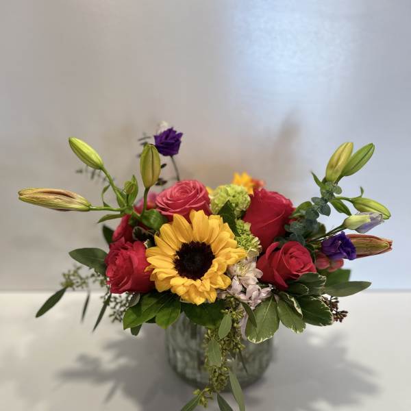 Mixed bouquet with a sunflower, pink roses, and purple blooms in a glass vase