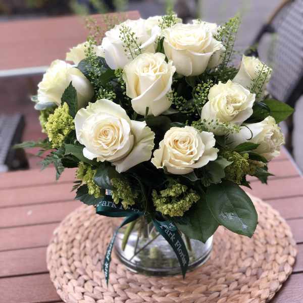 Round bouquet of white roses with green accents in a clear glass vase.