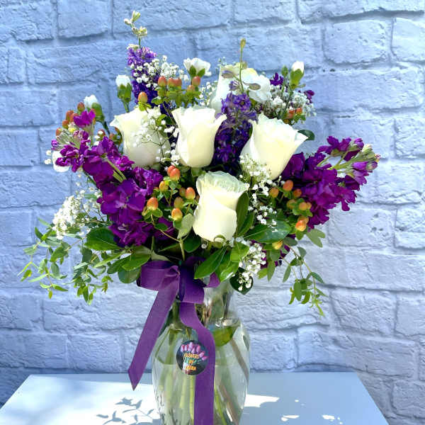 White roses and purple flowers arranged in a glass vase with a purple ribbon.