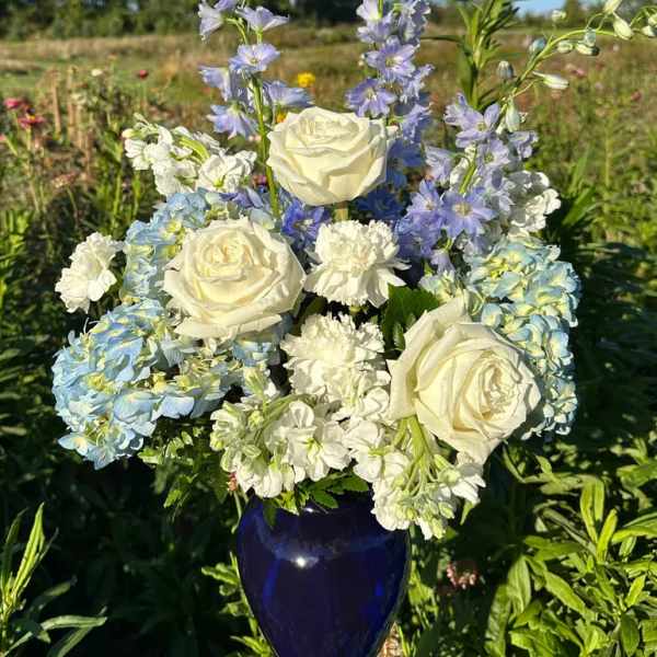 White roses, carnations, and blue hydrangeas with blue delphinium in a dark blue vase outdoors.
