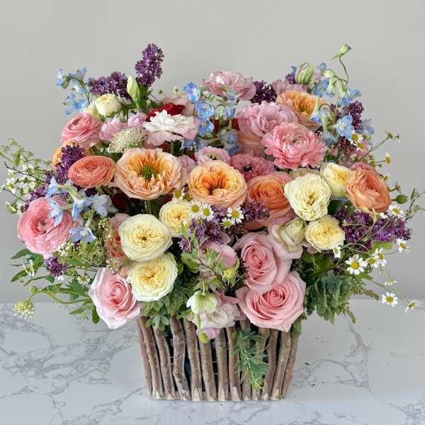 Lush arrangement of pink, peach and yellow roses with lilac and blue blooms in a rustic twig container