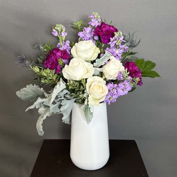 White roses and purple flowers arranged in a tall white vase.