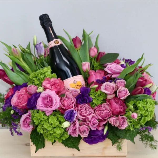 Wooden box arrangement of pink and purple flowers with a champagne bottle nestled in the center