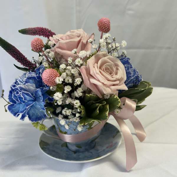 Pink roses and blue carnations arranged in a teacup with a ribbon
