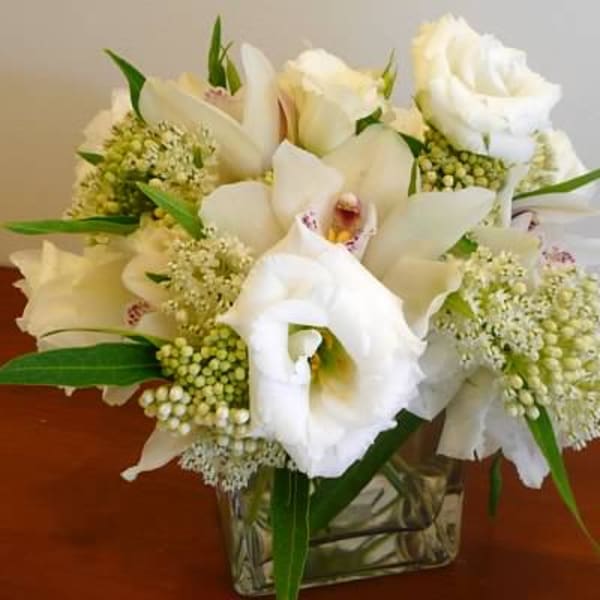 Low arrangement of white roses, lisianthus, and orchids in a clear glass cube vase
