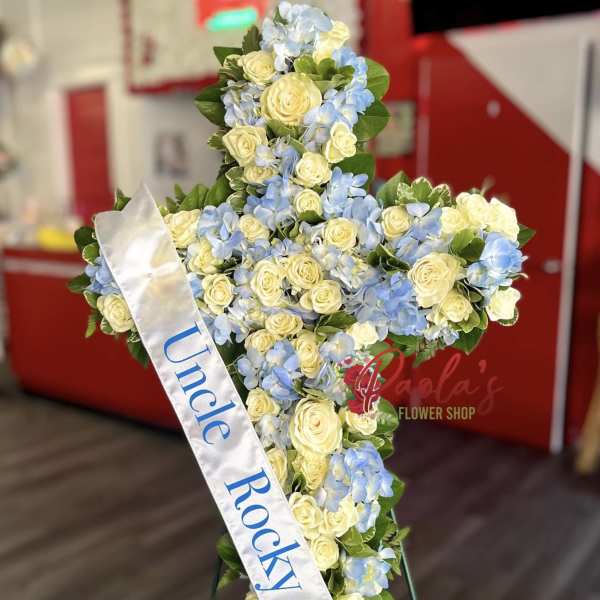 Cross-shaped floral tribute with pale roses and blue hydrangeas, draped with a white ribbon.