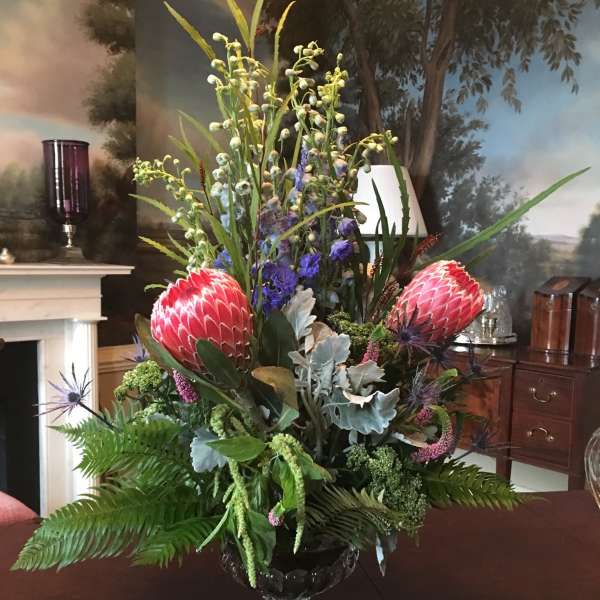 Tall tropical floral arrangement in a clear glass vase with pink protea and blue accents