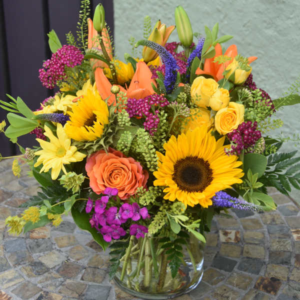Bright mixed arrangement of sunflowers, roses, lilies and daisies in a clear glass vase