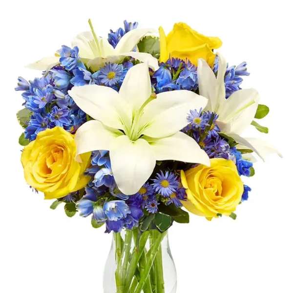 Clear vase of white lilies, yellow roses, and blue flowers in a rounded mixed bouquet