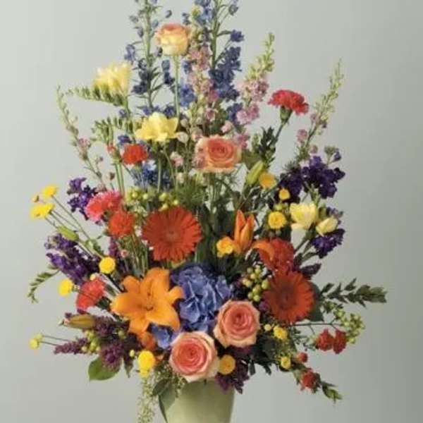 Tall mixed flower arrangement in a pale vase with orange, pink, yellow, and blue blooms