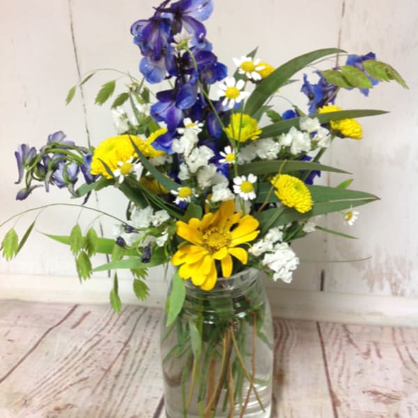 Wildflower-style bouquet of blue delphinium, yellow daisies, and white blooms in a clear glass jar vase