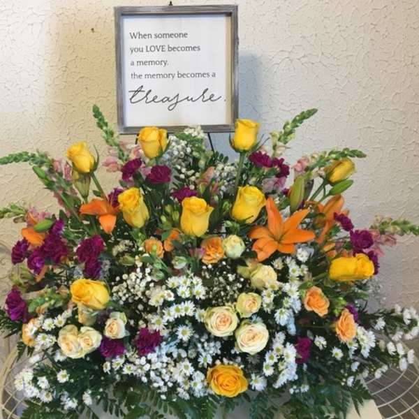 Large sympathy arrangement with yellow, orange, and purple flowers beneath a memorial sign