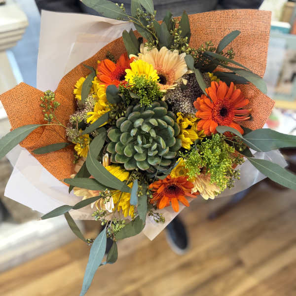 Hand-tied bouquet with a central green succulent and orange and yellow gerbera daisies in orange wrap