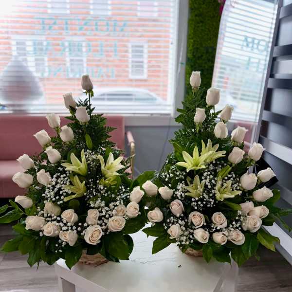 Pair of white rose and lily arrangements with baby's breath in woven baskets on a white table.