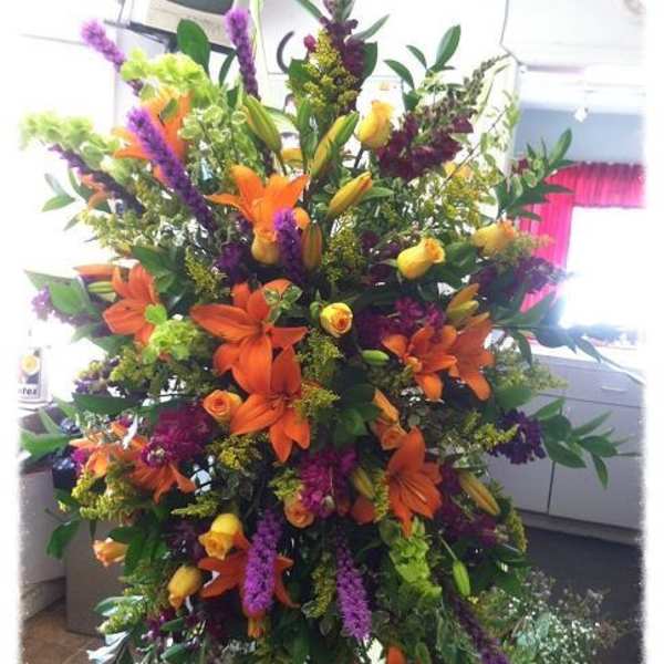 Tall standing spray of orange lilies, yellow roses, and purple flowers on a metal easel