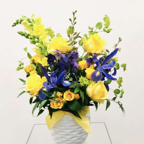 Yellow roses and blue irises in a white vase with a yellow ribbon
