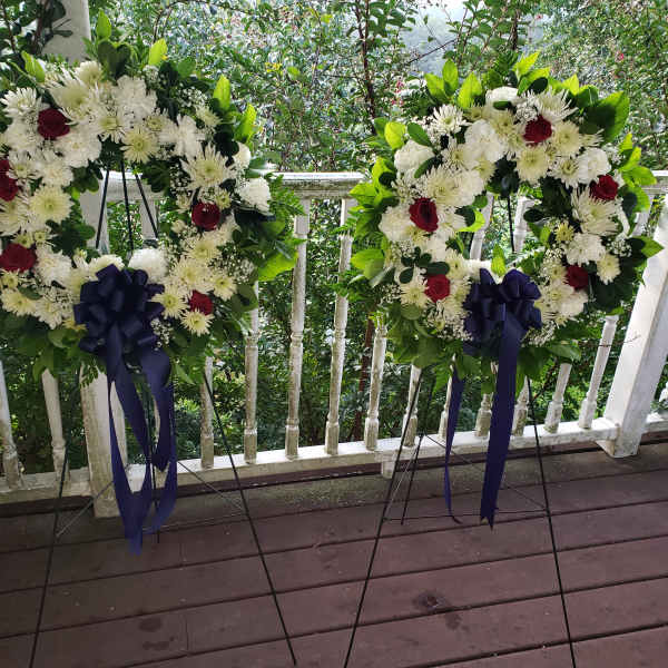 Two floral wreaths on stands with white flowers, red roses, and navy ribbons