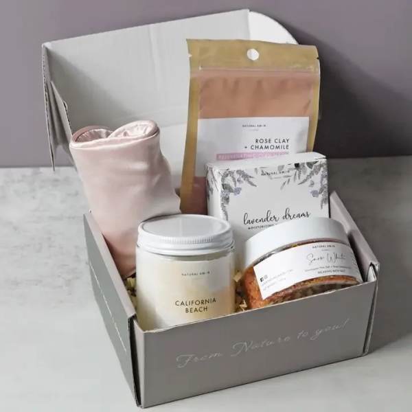 Open gray gift box with spa items including jars, boxed products, and a pink fabric accessory.