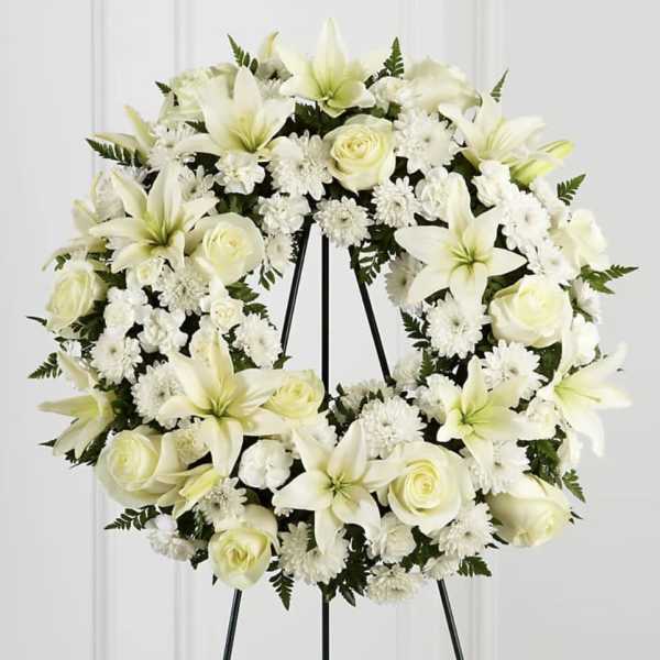 White floral wreath with lilies, roses, and daisies on a black stand