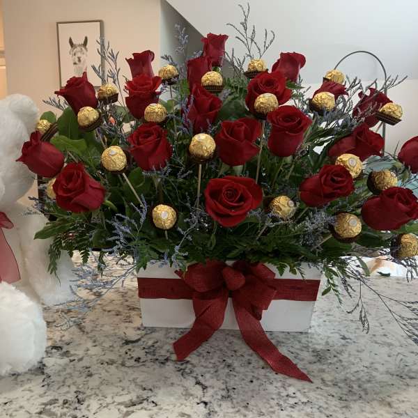 Red roses in a white box beside a white teddy bear with a red ribbon