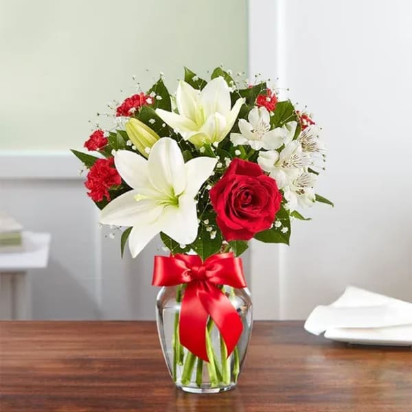 Bouquet of white lilies, red roses, and red carnations in a glass vase with a red ribbon.