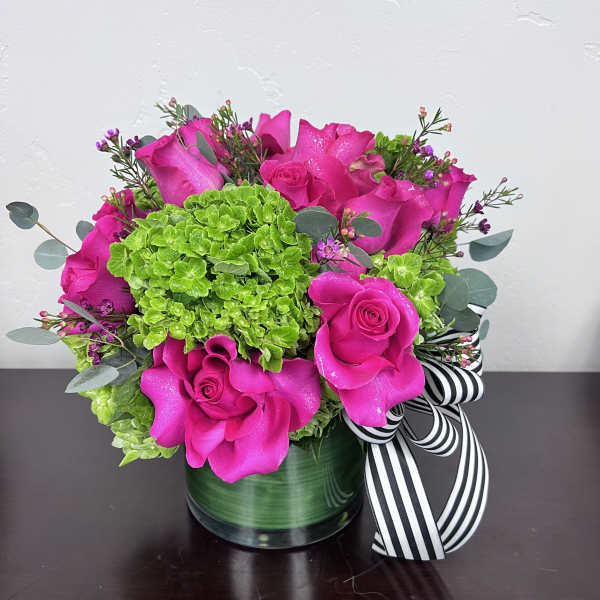 Compact arrangement of hot pink roses and green hydrangeas in a short glass vase with a striped ribbon bow.