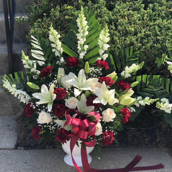 White lilies and red carnations arranged in a white urn with a pink ribbon