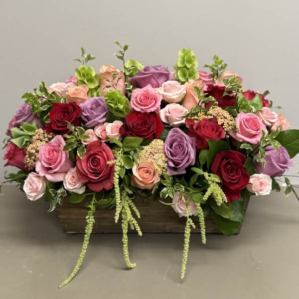 Low wooden box filled with red, pink, peach, and lavender roses with green accents.