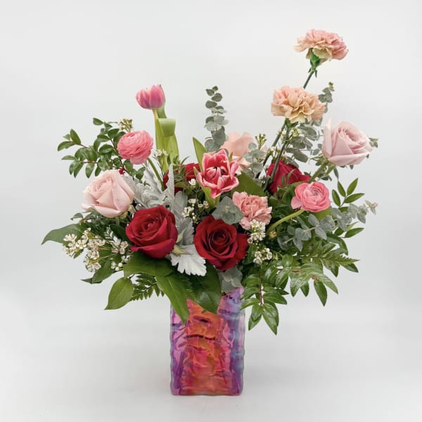 Mixed pink and red roses, tulips, ranunculus, and carnations in a colorful glass vase