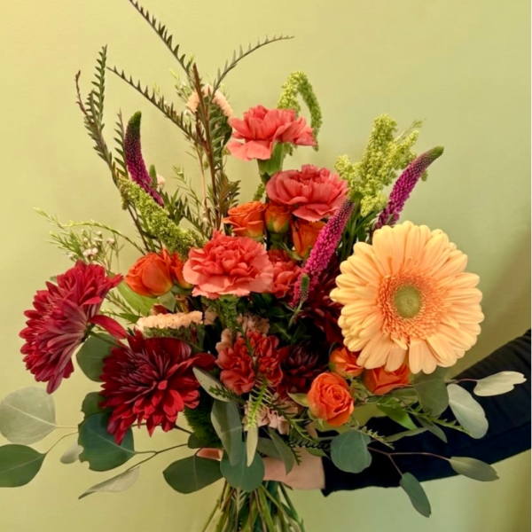 Hand-tied bouquet of orange gerbera, red mums, carnations, and roses with tall filler stems, held against a pale wall