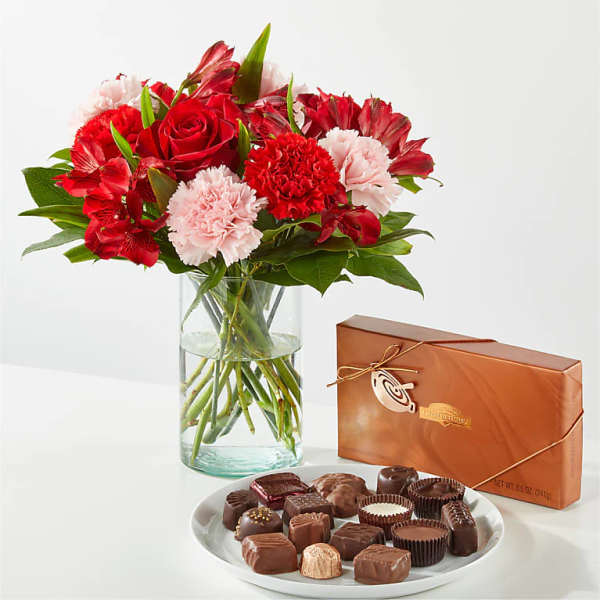 Red and pink flower arrangement in a glass vase with a box and plate of assorted chocolates