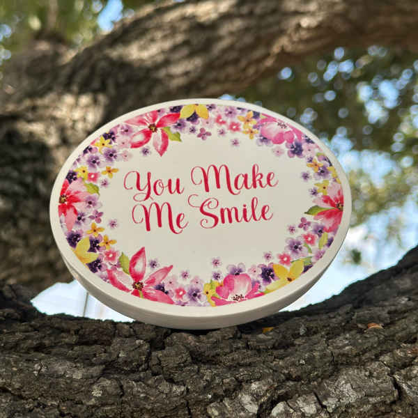 Oval floral sign reading "You Make Me Smile" on a tree branch