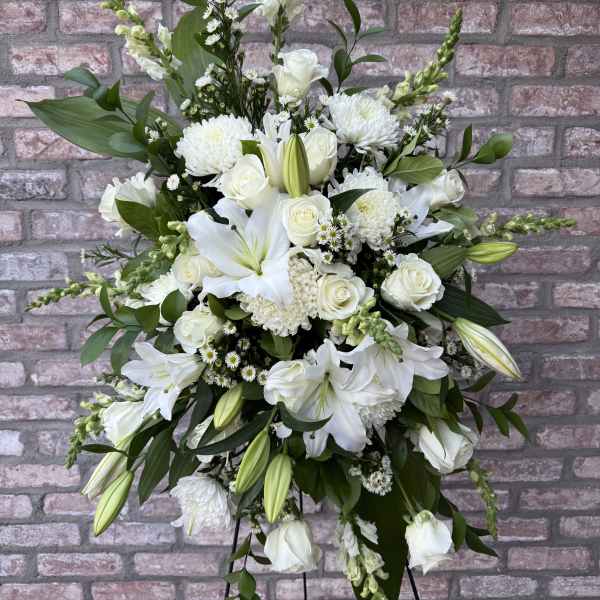 Tall all-white standing spray with lilies, roses, and mums on a metal easel against a brick wall.