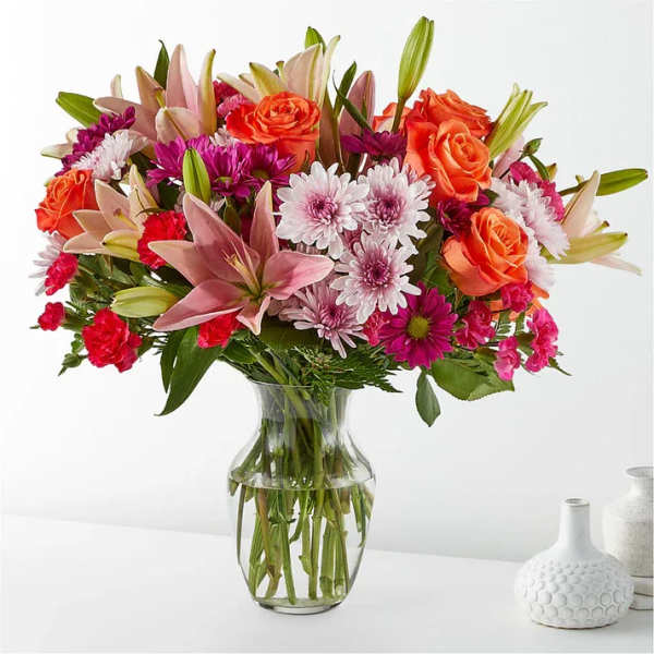 Mixed bouquet of orange roses, lilies, and pink daisies in a glass vase