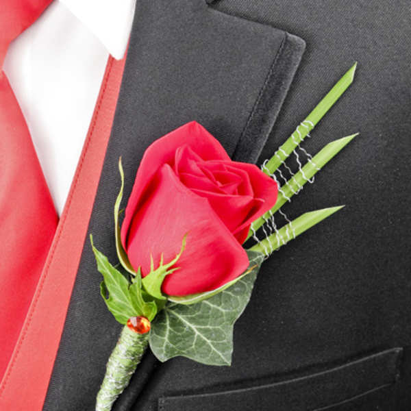 Single red rose boutonniere with decorative green blades on a black suit with red vest.