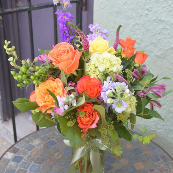 Mixed bouquet of orange roses, yellow blooms, and purple flowers in a clear glass vase