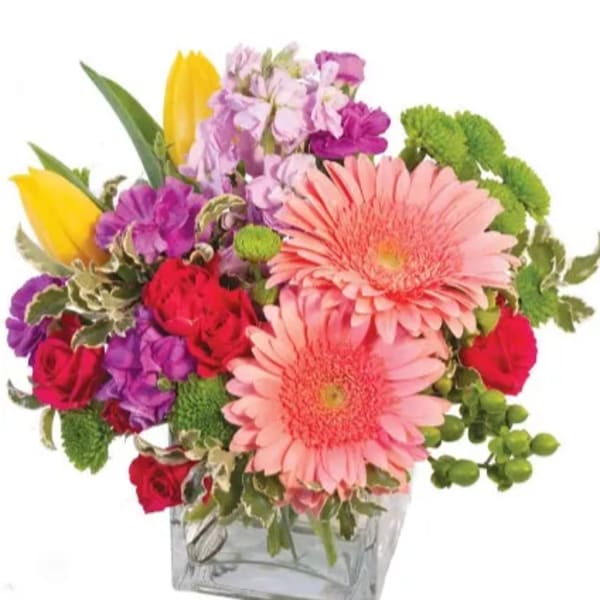 Colorful mixed bouquet with pink gerbera daisies, red roses, purple blooms, and yellow tulips in a glass cube vase