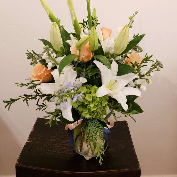 Tall arrangement of white lilies and peach roses with green hydrangea in a clear glass vase