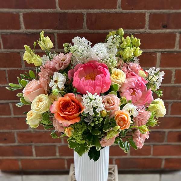 Tall arrangement of pink peonies, peach and orange roses, and white blooms in a white ribbed vase