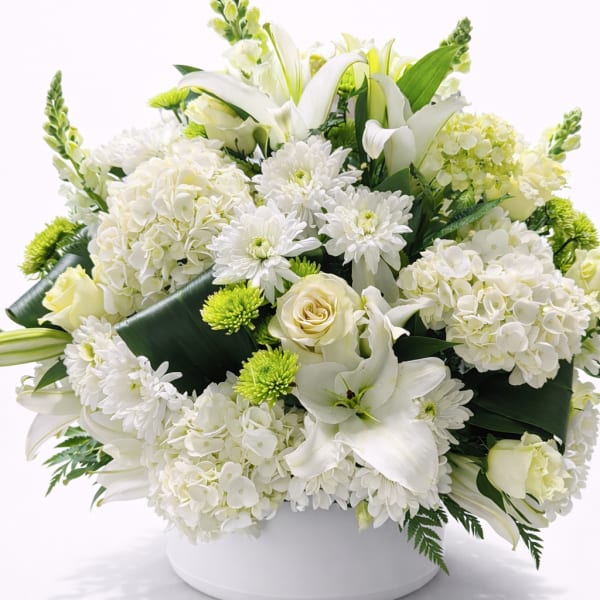 Low round arrangement of white lilies, roses, hydrangeas and green mums in a white bowl.