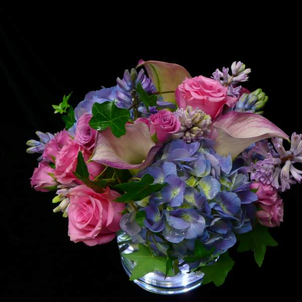 Low arrangement of pink roses, blue hydrangeas, and pale calla lilies in a clear glass vase.