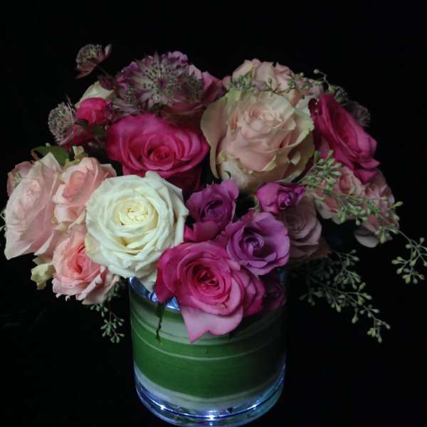 Compact arrangement of pink, peach, and cream roses in a clear glass vase with a green leaf wrap.