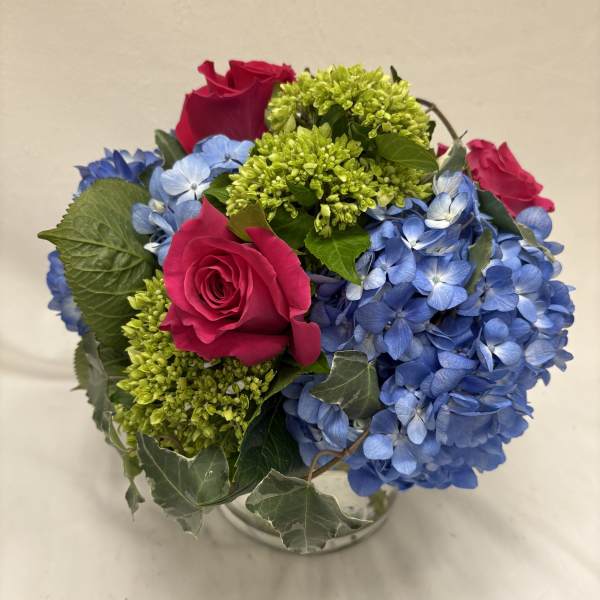 Compact arrangement of hot pink roses, blue hydrangeas, and green blooms in a clear glass vase with ivy.