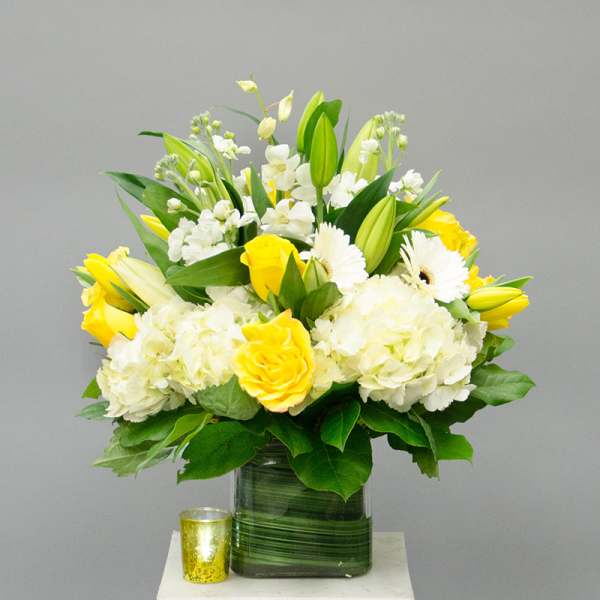 Yellow and white floral arrangement in a glass vase with a small candle beside it.