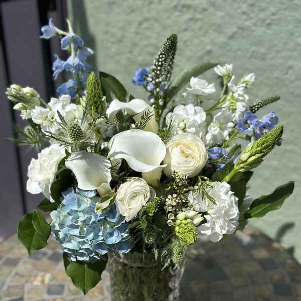 Arrangement of white calla lilies, roses, and blue hydrangea in a tall clear glass vase